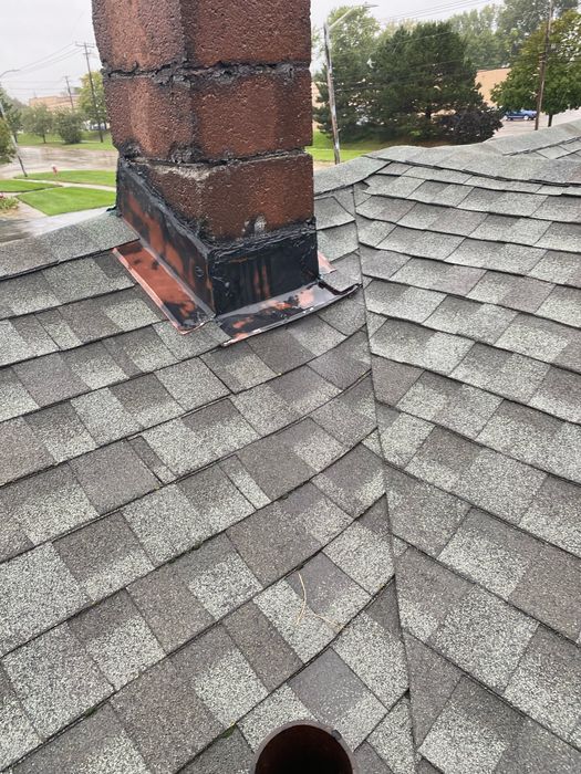 What we found before repair: a brick chimney on a gray asphalt shingle roof with the copper-colored flashing at its base smeared in black tar-like sealant instead of properly sealed, above a shingled valley, on an overcast day.