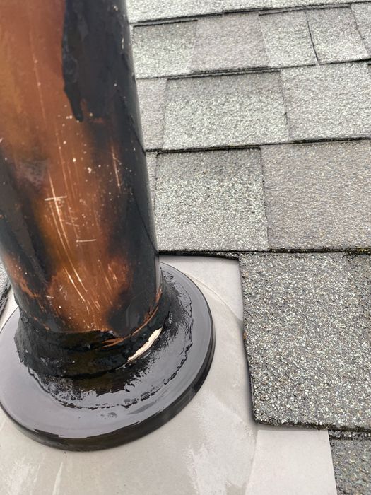 What we found before repair: close-up of a copper vent pipe through a gray pipe boot on asphalt shingles, with the black sealant at the collar cracked and pulled away from the pipe, leaving a visible gap.