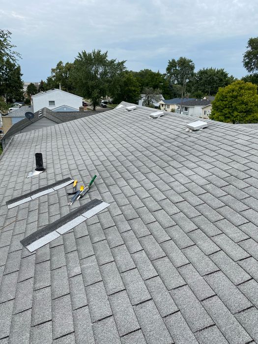 A real roof at the start of an inspection and repair visit: gray asphalt shingles with box vents near the ridge, a vent pipe, and replacement shingle pieces and hand tools laid out before the work was done.