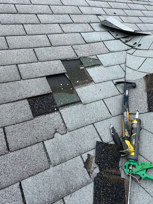 What we found before repair: gray asphalt shingle roof with missing and torn shingles exposing underlayment and nail heads, a lifted shingle tab farther up the slope, and a hammer, utility knife and caulk gun set down as the repair was starting.